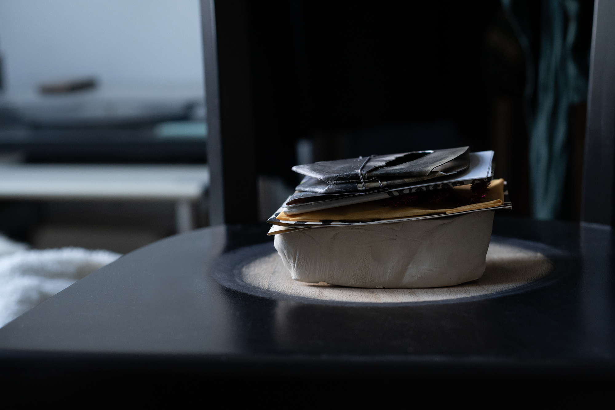 The picture is a photograph of a pile of diverse materials whose exact content can only be adumbrated because it must be unfolded to be seen. The colors of the materials are yellow, and lack, purple. The overall atmosphere of the picture is ghosty and calm. The materials are placed on a piece of dried clay.